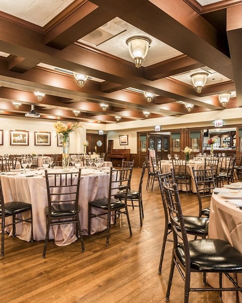 Hugos-Chicago-Private-Dining-1 Elegant banquet hall with round tables draped in white cloth, surrounded by dark wooden chairs. Soft lighting and floral centerpieces create a warm ambiance.